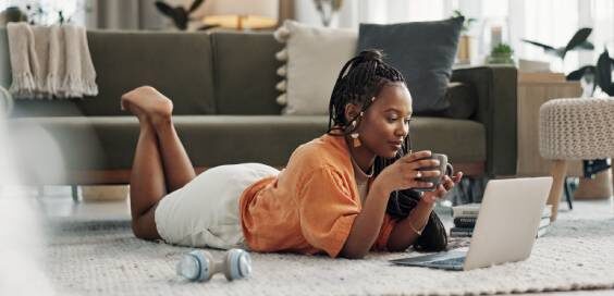 A young woman lies on her living room rug, holding a coffee mug, looking at her laptop. Books and headphones are by her side.