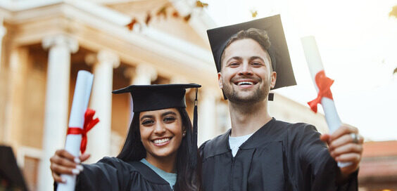 Two graduate students in their cap and gowns, holding their graduate degrees while posing for a photo at their school.