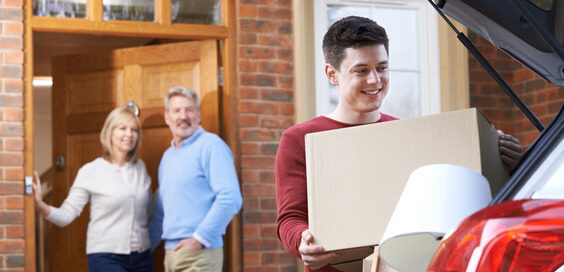 A young man smiles as he loads boxes into the back of a car. His parents smile and stand by the front door.
