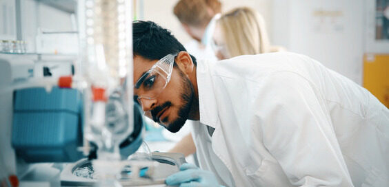 Young college student in laboratory carefully examining an experiment. He's wearing proper PPE including gloves and safety goggles.