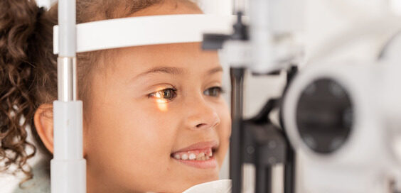 A smiling girl with curly pigtails resting her chin on a chin rest during an eye exam in a doctor's office.