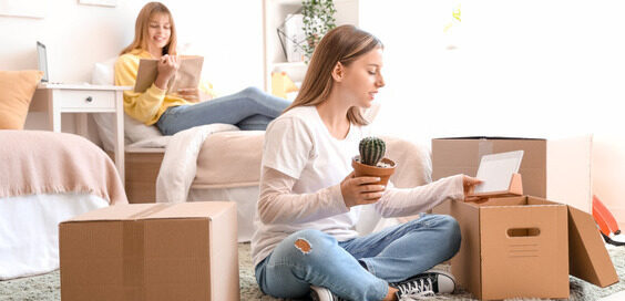 A young woman sitting on the floor of a bright room with cardboard boxes and another woman reading on a bed behind her.