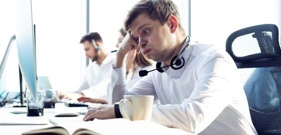A man with a headset sits at a long desk with coworkers, resting his head on his hand, looking stressed.