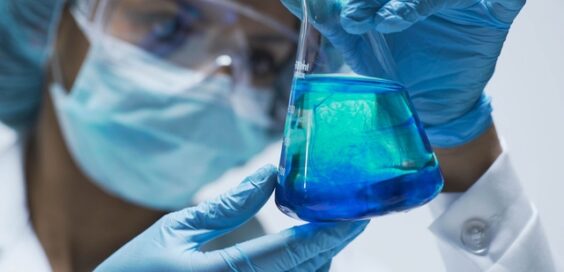 A woman wearing blue latex gloves, a face mask, goggles, and a scrub cap holding a beaker filled with blue liquid.