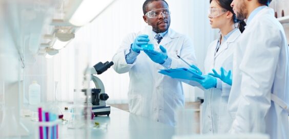 Two men and one woman stand by a white counter with a microscope and test tubes; one man holds a flask with blue liquid.