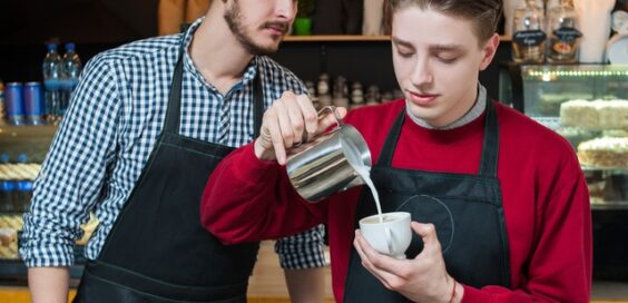 A young person in a black apron carefully pours milk into a small cappuccino cup while another barista observes them.