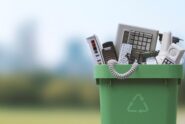 A close-up view shows a green bucket with the recycling symbol, filled with phones, remotes, and calculators.