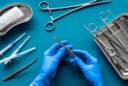 A doctor wearing blue gloves holds a scalpel. On the table are other surgical tools, including needles and forceps.