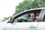 A young man smiling and looking out the window while sitting in the driver’s side seat of a car during the day.
