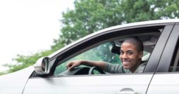 A young man smiling and looking out the window while sitting in the driver’s side seat of a car during the day.