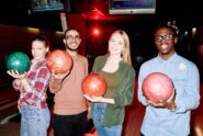 Four smiling young adults standing inside a bowling alley with colorful bowling balls in their hands.