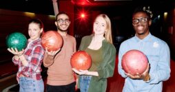 Four smiling young adults standing inside a bowling alley with colorful bowling balls in their hands.