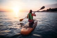 A woman in an orange shirt and green life vest sitting on a paddleboard as the sun goes down in front of her.
