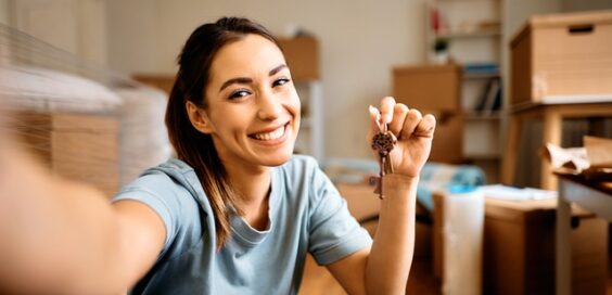 A young woman taking a selfie with the keys to her new apartment. Moving boxes fill the background.