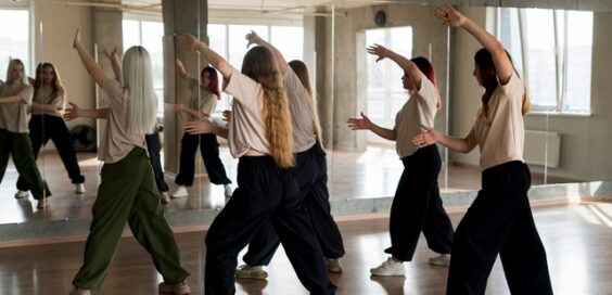 A group of young female dancers practices in their studio, all wearing matching white shirts, in front of a large mirror.