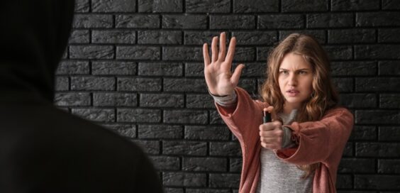 A young woman stands against a black brick wall, a shadow man in front of her. She holds out her hand and pepper spray.