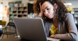 A college student leans her head against her hand as she stares at her computer screen, a blank expression on her face.