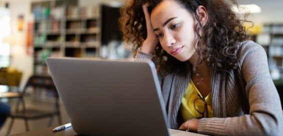 A college student leans her head against her hand as she stares at her computer screen, a blank expression on her face.