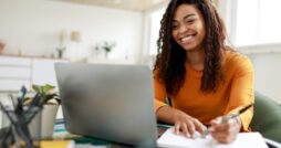 A young Black woman smiles as she sits at a desk, using a personal computer and writing in her notebook.