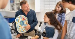 An educator points to a plastic model of a cell in front of a group of young students intently listening to him.