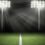 Two sets of tall lighting towers overlook and illuminate the green grass of a football stadium with a white stripe.