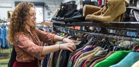 A woman browsing through shirts that are hanging on a rack. There are multiple pairs of shoes on top of the rack.