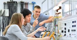 A group of students using meters to measure electrical current from wires leading to illuminated bulbs in a lab space.