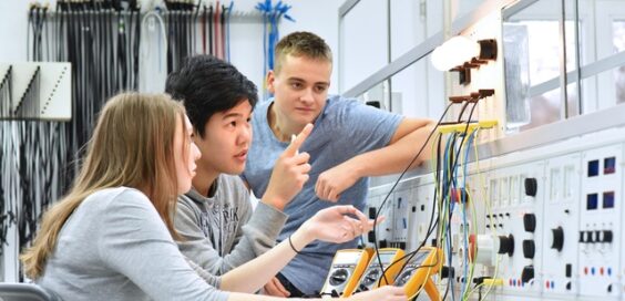 A group of students using meters to measure electrical current from wires leading to illuminated bulbs in a lab space.