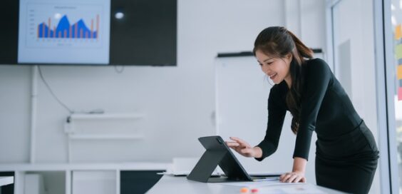 A woman stands at a table with a tablet open and propped up in front of her. Behind her is a TV with graphs on it.