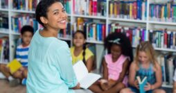 A young person sitting in a library area with students and looking over their shoulder to smile toward the viewer.