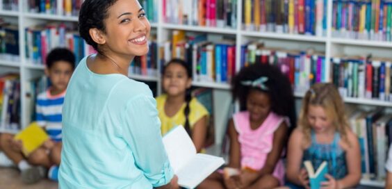 A young person sitting in a library area with students and looking over their shoulder to smile toward the viewer.