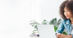 A young woman with curly hair is sitting in a white room, studying at a laptop with notebooks and a calculator.