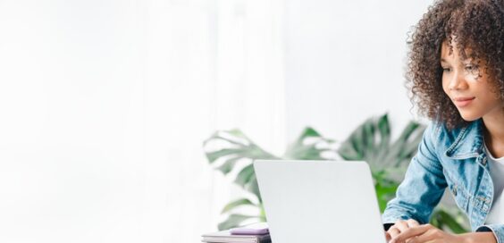 A young woman with curly hair is sitting in a white room, studying at a laptop with notebooks and a calculator.