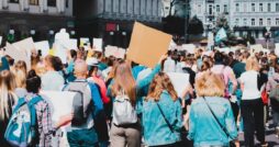 A crowd at a city protest, seen from behind, holding blank signs with urban buildings and trees in the background.