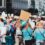 A crowd at a city protest, seen from behind, holding blank signs with urban buildings and trees in the background.