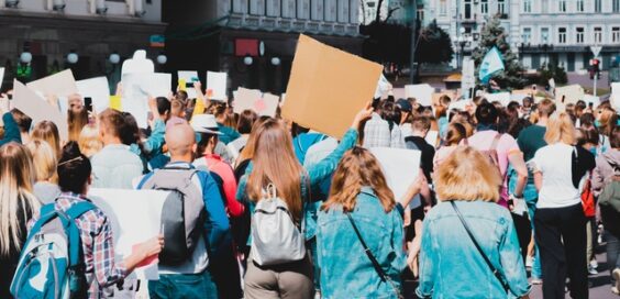 A crowd at a city protest, seen from behind, holding blank signs with urban buildings and trees in the background.