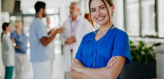 A young woman wearing blue scrubs stands in a hospital corridor and smiles with her arms folded across her chest.