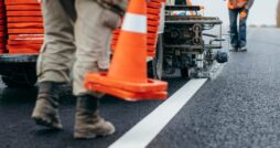 A close-up of two street workers laying orange cones down as the truck paints white lines along the roadway.