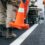 A close-up of two street workers laying orange cones down as the truck paints white lines along the roadway.