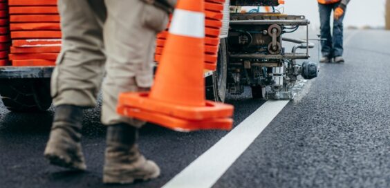 A close-up of two street workers laying orange cones down as the truck paints white lines along the roadway.