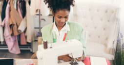 A young woman at a sewing machine has a measuring tape around her neck. Garments hang on a clothes rack behind her.