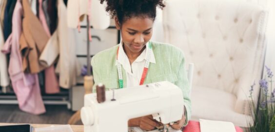 A young woman at a sewing machine has a measuring tape around her neck. Garments hang on a clothes rack behind her.