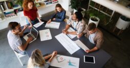Team of coworkers collaborating around a table reviewing charts, documents, and digital devices during a business meeting in a modern office