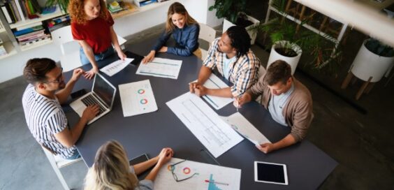 Team of coworkers collaborating around a table reviewing charts, documents, and digital devices during a business meeting in a modern office