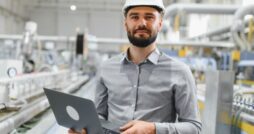 A man in a white hard hat holds a laptop on a production floor, with blurred machinery behind him.