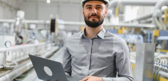 A man in a white hard hat holds a laptop on a production floor, with blurred machinery behind him.