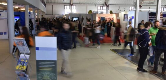 A crowded indoor professional event with attendees walking past booths, signage, and a brochure display stand.