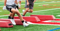 A close-up of the feet of three young athletes navigating around red barriers on a football field for a drill.