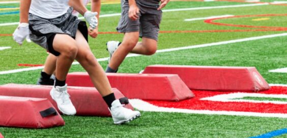 A close-up of the feet of three young athletes navigating around red barriers on a football field for a drill.