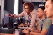 Instructor in glasses leans over student at computer, pointing at screen in classroom with other learners seated nearby.
